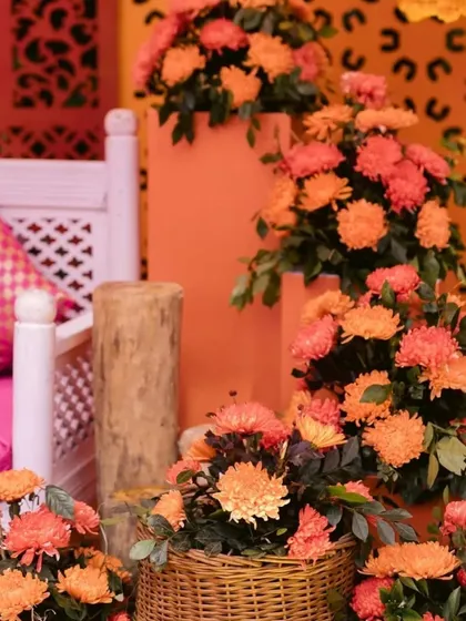 A close-up of orange floral arrangements for a mehendi ceremony. The flowers are placed in wicker baskets and on wooden logs, adding a rustic and natural touch to the vibrant decor.