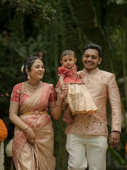A full-length view of a family in their coordinated ethnic wear. The father's peach jacket features subtle embroidery that echoes the colors in the mother's saree and the child's traditional dress.