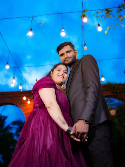 A romantic pre-wedding photo under a canopy of hanging lights. The bride's purple gown and the beautiful twilight sky create a magical atmosphere.