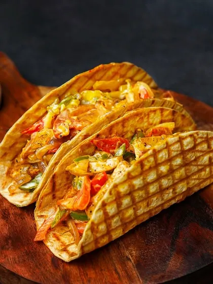 Grilled tacos filled with chicken and colorful bell peppers, served with a side of sauce. The warm tones of the food are enhanced by the dark, rustic background.