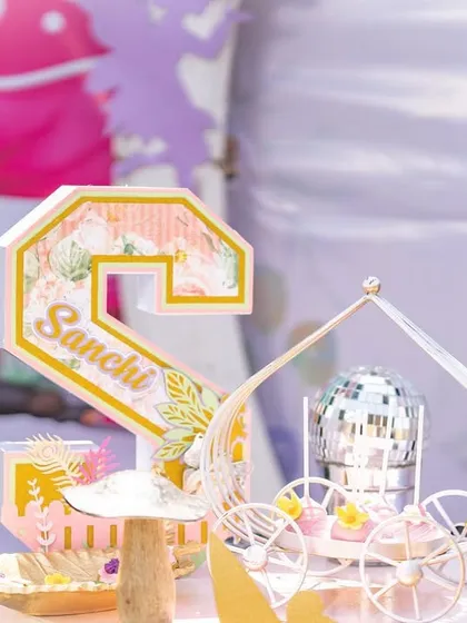 A personalized 'Sanchi' name cutout for a second birthday, designed with a floral pattern and placed next to a miniature carriage holding a disco ball.