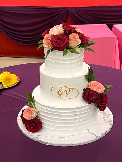 A classic three-tier white wedding cake with a simple, elegant ridged texture. It's decorated with a cascade of fresh red and peach roses and features a custom gold monogram, proving that true beauty lies in simplicity.