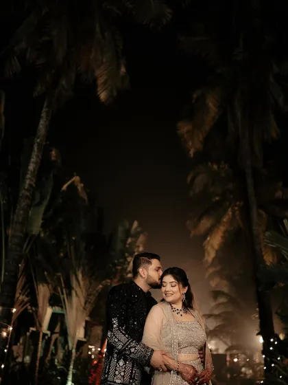 A romantic nighttime portrait under the palm trees. The groom's gentle kiss on the bride's forehead, illuminated by soft lights, creates a magical and intimate moment.