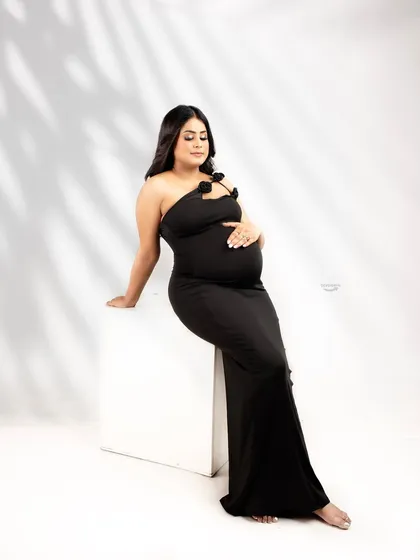 A modern and chic solo portrait. The client wears a simple, elegant black gown, seated against a clean white background with artistic shadow play for a contemporary maternity look.