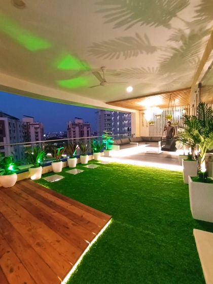 A stunning view of a large balcony makeover in Rajkot at dusk. The combination of artificial grass, a wooden deck, and creative ceiling light projections creates a magical atmosphere.