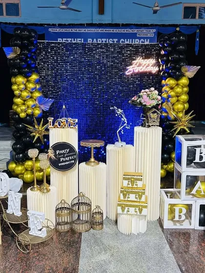 This large-scale birthday decoration features a stunning blue shimmer wall as the centerpiece. The wall is framed by black and gold balloon columns, multiple pedestals, and 'BABY' blocks, creating a grand display.