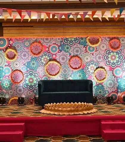 A vibrant and colorful stage backdrop for a Haldi function. The design uses a mandala-printed flex and decorative hangings to create a festive and energetic photo corner.