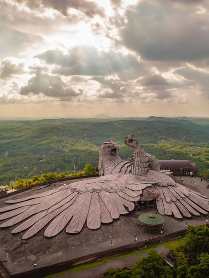 A stunning aerial photograph of the Jatayu Earth's Center in Kerala, featuring the world's largest bird sculpture. The divine light breaking through the clouds adds a mythical quality to the scene.