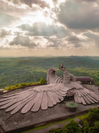 A stunning aerial photograph of the Jatayu Earth's Center in Kerala, featuring the world's largest bird sculpture. The divine light breaking through the clouds adds a mythical quality to the scene.