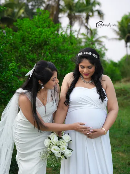 A tender moment between friends, with one gently touching the baby bump. Both are dressed in elegant white gowns.