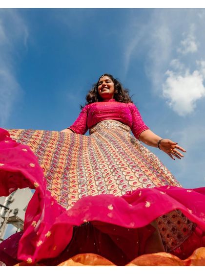 A low-angle shot capturing the bride's vibrant personality and the beautiful flare of her lehenga against a bright blue sky. A moment of pure, uninhibited joy.