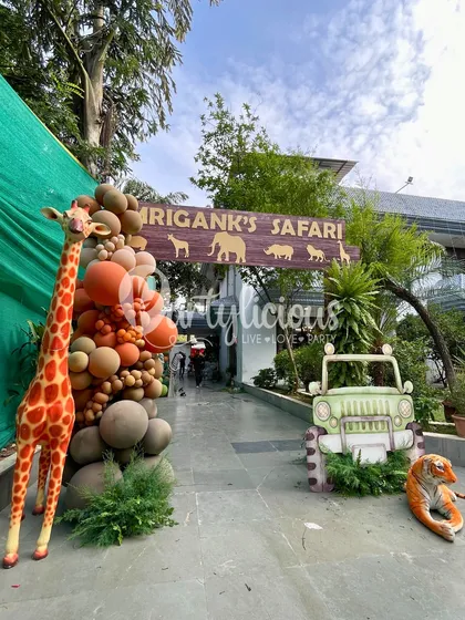 The grand entrance to Mrigank's Safari, featuring a custom sign, a friendly tiger, a safari jeep, and a towering giraffe to welcome guests.