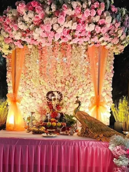A minimalist yet beautiful design featuring a wall of pink and white flowers with a decorative peacock. The soft orange drapes and lighting create a warm and inviting space for Bappa.
