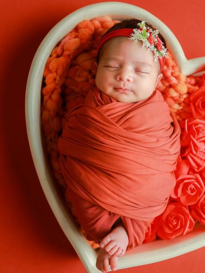 A close-up of a baby sleeping in a heart-shaped bowl. The rich red color of the wrap and roses creates a feeling of warmth and love.