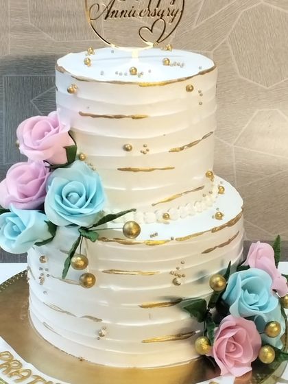 A classic two-tiered anniversary cake with a textured buttercream finish, gold accents, and a mix of pink and blue sugar roses.
