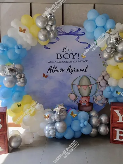 Announce the arrival of your little prince with this adorable "It's a Boy!" baby announcement decoration. The setup features a custom circular backdrop with a hot air balloon theme, surrounded by a balloon arch in shades of blue, yellow, and silver.