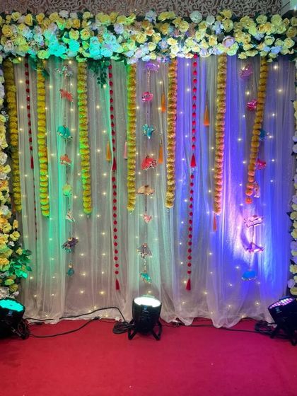 A complete stage frame for a Haldi function, covered in yellow flowers. The backdrop includes a sheer curtain with fairy lights and hanging marigold strings for a classic festive feel.
