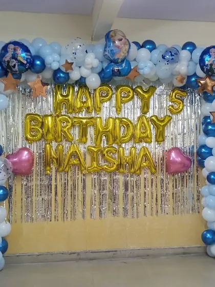 A Frozen-themed backdrop for a 5th birthday. This setup includes a balloon arch in blue and white with character balloons, all framing a silver tinsel curtain and 'Happy Birthday' foil letters.