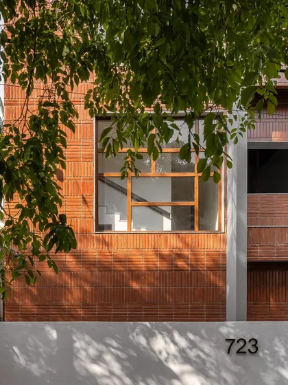 A close-up of the facade, showing the interplay between the warm brick, the smooth white plaster, and the large wooden window frame. The natural foliage adds a soft contrast to the building's clean lines.