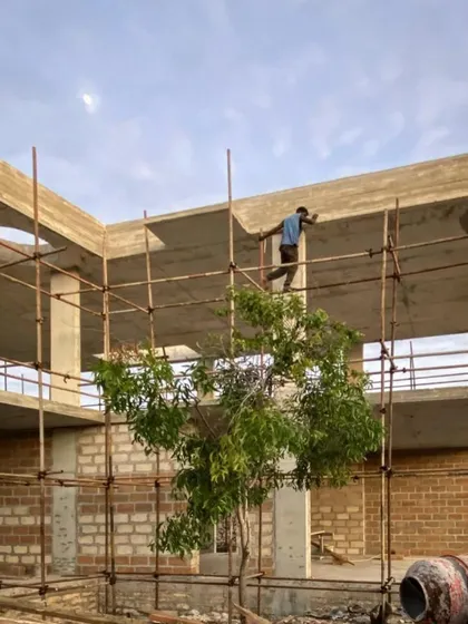 The concrete frame of a phase two building at Kizikula is completed. A worker walks along the scaffolding against the sky, highlighting the verticality of the structure and the open-air design.