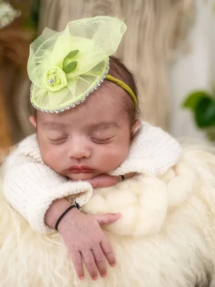 A close-up on the baby's peaceful face, highlighting the delicate features and the cute lime green fascinator. The soft, furry texture below adds to the cozy, natural vibe.