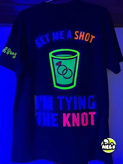The back of the bachelor party T-shirt under blacklight. The neon colors of the "I'm tying the knot" design shine brightly.