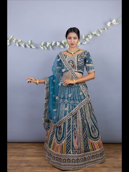 A full-length view of the teal lehenga. The skirt features intricate embroidery with pops of pink and orange, and the sheer dupatta adds a layer of elegance.
