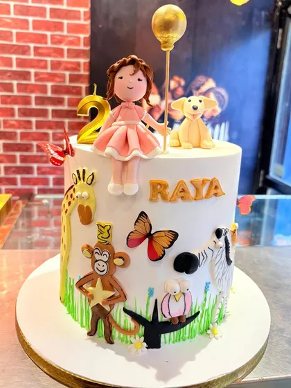 A whimsical animal-themed cake for Raya's 2nd birthday, featuring a fondant doll, a dog, and various jungle animals.