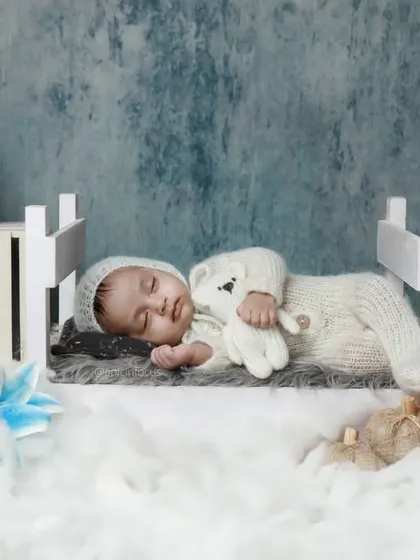 A baby in a white knit outfit sleeping on a miniature bed, set against a cool blue backdrop with matching floral accents.