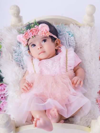 A close-up portrait of a beautiful baby girl wearing a pink dress and a floral crown. Her curious expression is captured perfectly in this sweet and gentle photograph.