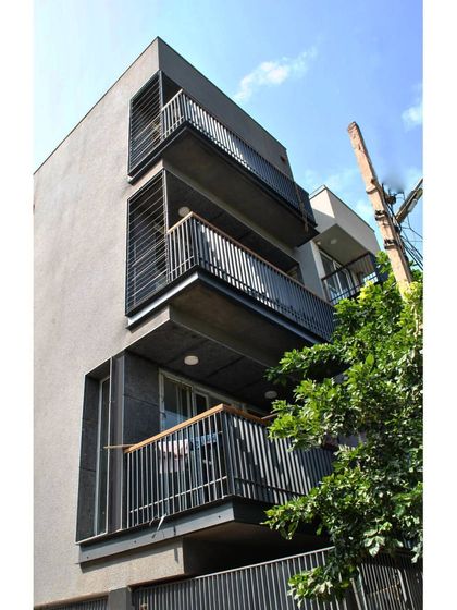 A corner view of a completed residence in Gurgaon. The design uses a simple grey facade punctuated by projecting steel balconies with wooden handrails, creating a clean, modern aesthetic for an urban apartment block.