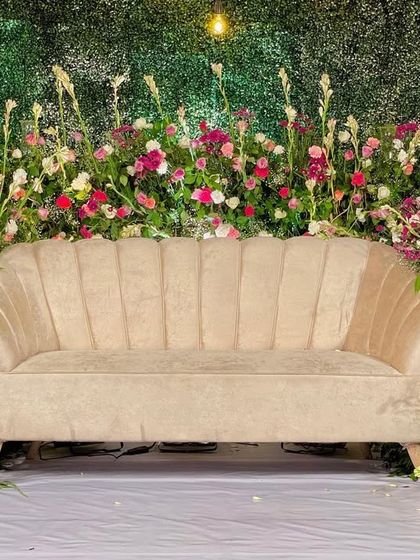 A modern photo booth backdrop. A simple yet elegant beige sofa is set against a wall of greenery, with a wildflower-style arrangement of pink, white, and red blooms creating a beautiful frame.