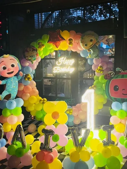 We love creating magical parties for kids, like this vibrant Cocomelon first birthday. The setup includes a colorful balloon arch, character cutouts of JJ and the watermelon TV, a neon sign, and a light-up number '1'.