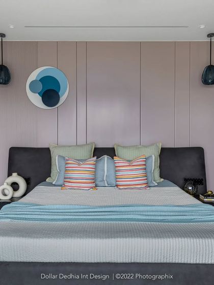 A full view of the pastel bedroom, with its lavender paneled wall, dark grey upholstered bed, and black accent lights that ground the soft color scheme.