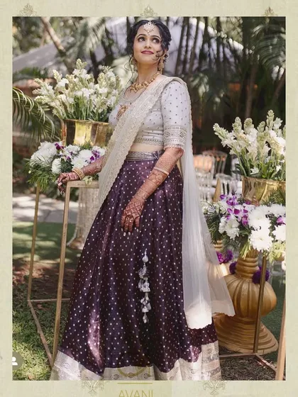 A full-length view of the bride in her purple polka-dot lehenga, standing in a beautiful garden setting. The outfit is perfect for a daytime wedding ceremony.