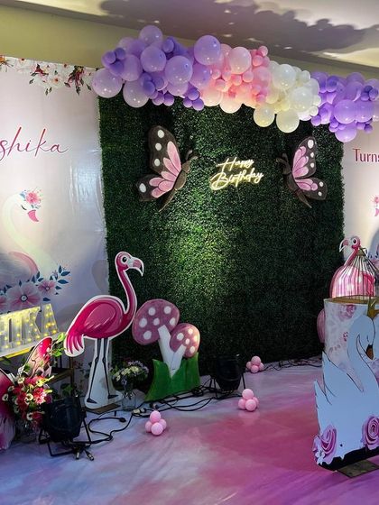 A whimsical garden theme combining butterflies, flamingos, and swans. The backdrop is a lush green wall with a neon sign, surrounded by pastel balloon garlands and cute props.