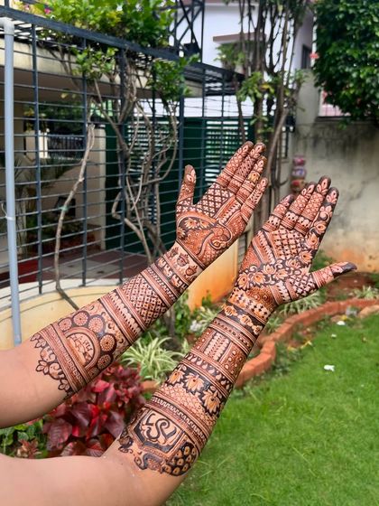The stained version of the bridal mehendi, showing the rich color against a garden background. The personalized "S" initial is visible on the arm.