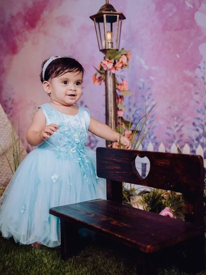 A baby girl in a lovely blue dress stands by a miniature park bench and lamppost. This themed setup shows the creative and detailed environments I build for baby photoshoots.