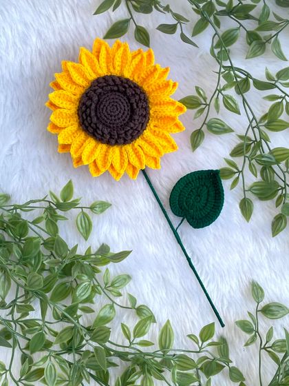 A beautiful, large crochet sunflower with a single leaf, lying on a soft white background. The vibrant yellow and rich brown center are so eye-catching.