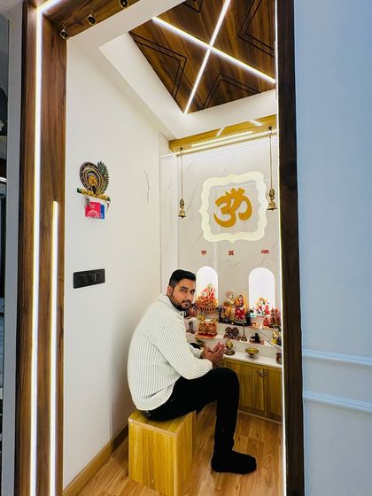 Sitting in the serene pooja room we designed at Prateek Laurel. The space features a custom backlit panel and a beautiful wooden ceiling design.