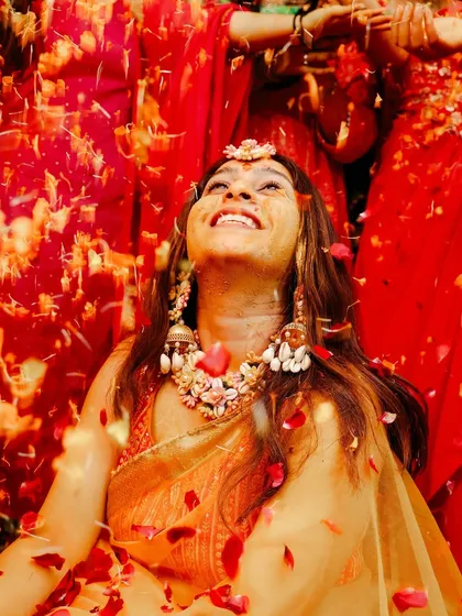 A moment of pure bliss. The bride looks up with a wide smile as she is showered with red flower petals during her Haldi ceremony.
