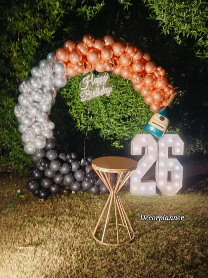 A daytime view of a stylish outdoor birthday setup. This image showcases a beautiful balloon arch in black, silver, and rose gold against a natural green backdrop, complete with '26' marquee numbers, perfect for a chic garden party.
