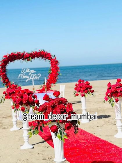 A wide-angle view of the beach marriage proposal setup, showing the red carpet aisle leading to the heart-shaped floral arch.