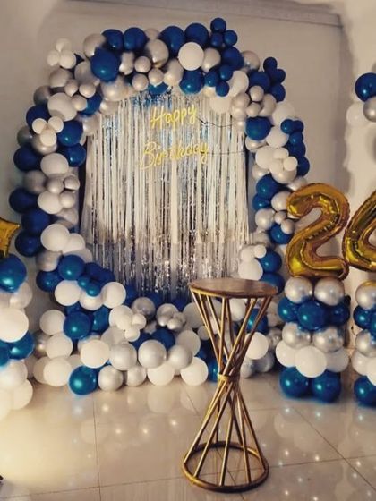 A vibrant 24th birthday decoration featuring a blue, white, and silver balloon arch over a silver foil fringe backdrop, complete with foil number balloons on pillars.