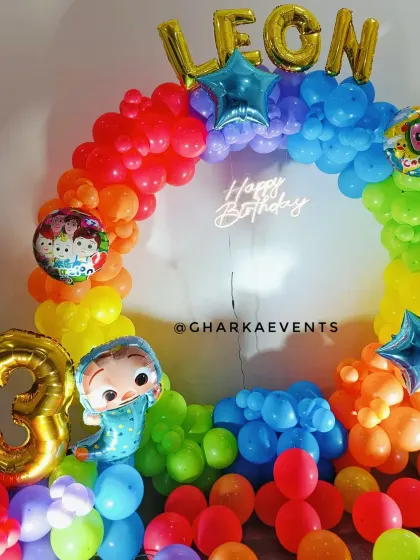 A vibrant rainbow Cocomelon-themed 3rd birthday. The circular balloon arch is filled with bright colors, Cocomelon character balloons, and a 'Happy Birthday' neon sign.