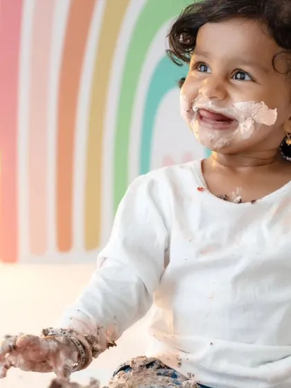 A close-up of a baby girl enjoying her cake smash, with a look of pure delight and a face covered in delicious frosting. The colorful rainbow backdrop adds to the cheerful atmosphere.