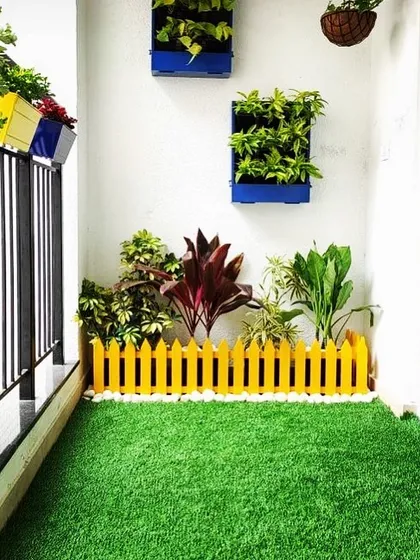 A bright and cheerful balcony floor. The entire space is covered in vibrant artificial grass, with a small yellow picket fence adding a playful touch and separating the potted plants.