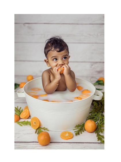 A delightful photo of a baby boy sitting in a miniature tub for a milk bath, surrounded by bright oranges. He is happily nibbling on an orange slice, creating a fun and vibrant image.