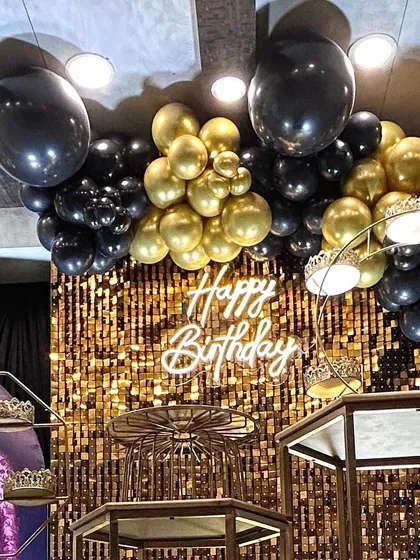 A chic and glamorous birthday celebration featuring a gold shimmer wall that catches the light beautifully. The setup is completed with a custom neon "Happy Birthday" sign and a dramatic black and gold balloon garland.