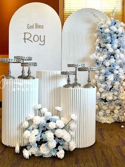 A unique baptism theme for a baby boy in shades of blue and white. The decor includes a custom backdrop, a floral tree, and textured plinths with silver cake stands for a modern and stylish look.